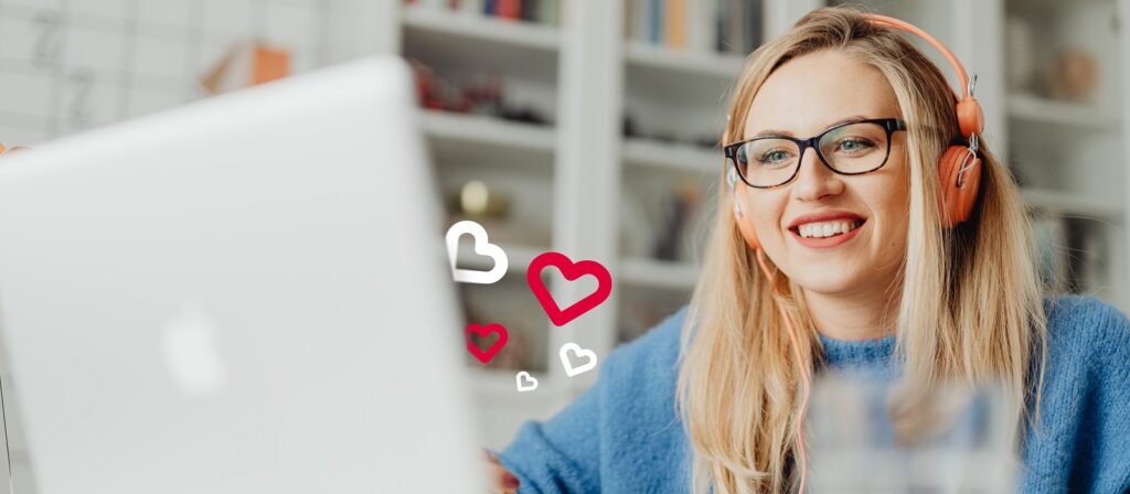 A woman wearing orange headphones and glasses smiles at a laptop screen, with illustrated heart icons floating near her. Bookshelves and a blurred background are visible behind her.