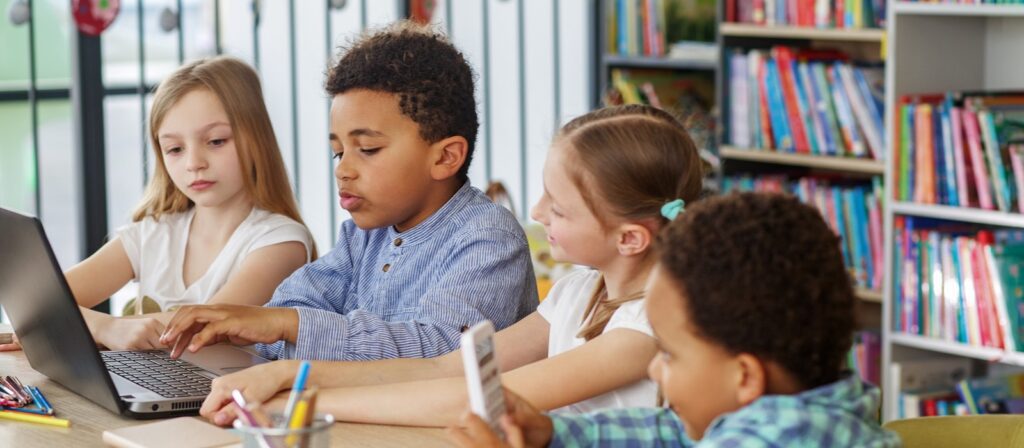 Four children sit at a table in a library or classroom. Two are using a laptop, one is holding a smartphone, and bookshelves are visible in the background. They appear to be working or studying together.