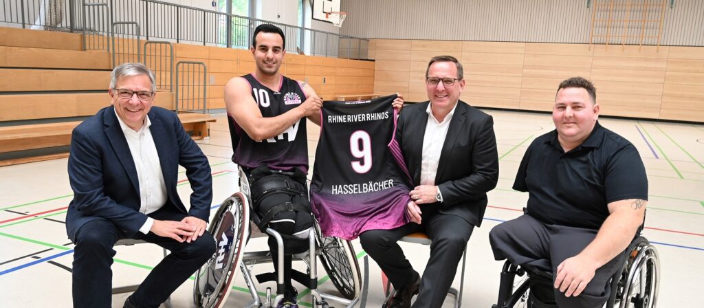 Four men, three in wheelchairs and one seated, pose together in a gym. One man in a basketball uniform holds up a team jersey with the name Hasselbachert and number 9. All are smiling for the photo.
