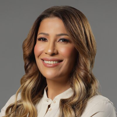A woman with long, wavy brown hair and blonde highlights smiles at the camera. She is wearing a light-colored blouse and posing against a plain, gray background.