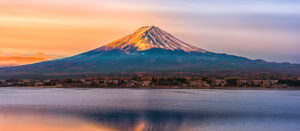 Mount Fuji at sunrise, its snow-capped peak glowing with warm colors, reflected in the calm water of a lake below. A small town sits between the mountain and the lake.