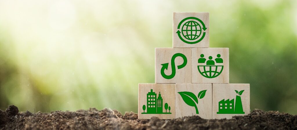 Wooden blocks stacked in a pyramid shape on soil, each block displaying green environmental icons such as a globe, leaf, group of people, buildings, recycling, and sustainable industry, with a blurred natural background.