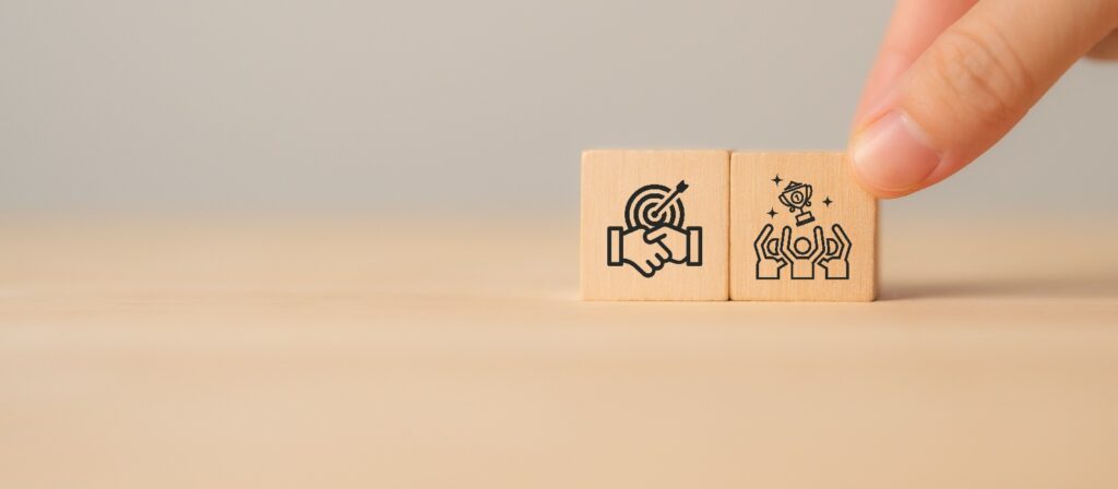 A hand arranges two wooden blocks. One block shows a handshake with a target, symbolizing partnership or goal achievement. The other shows raised hands with a trophy, representing celebration or success.