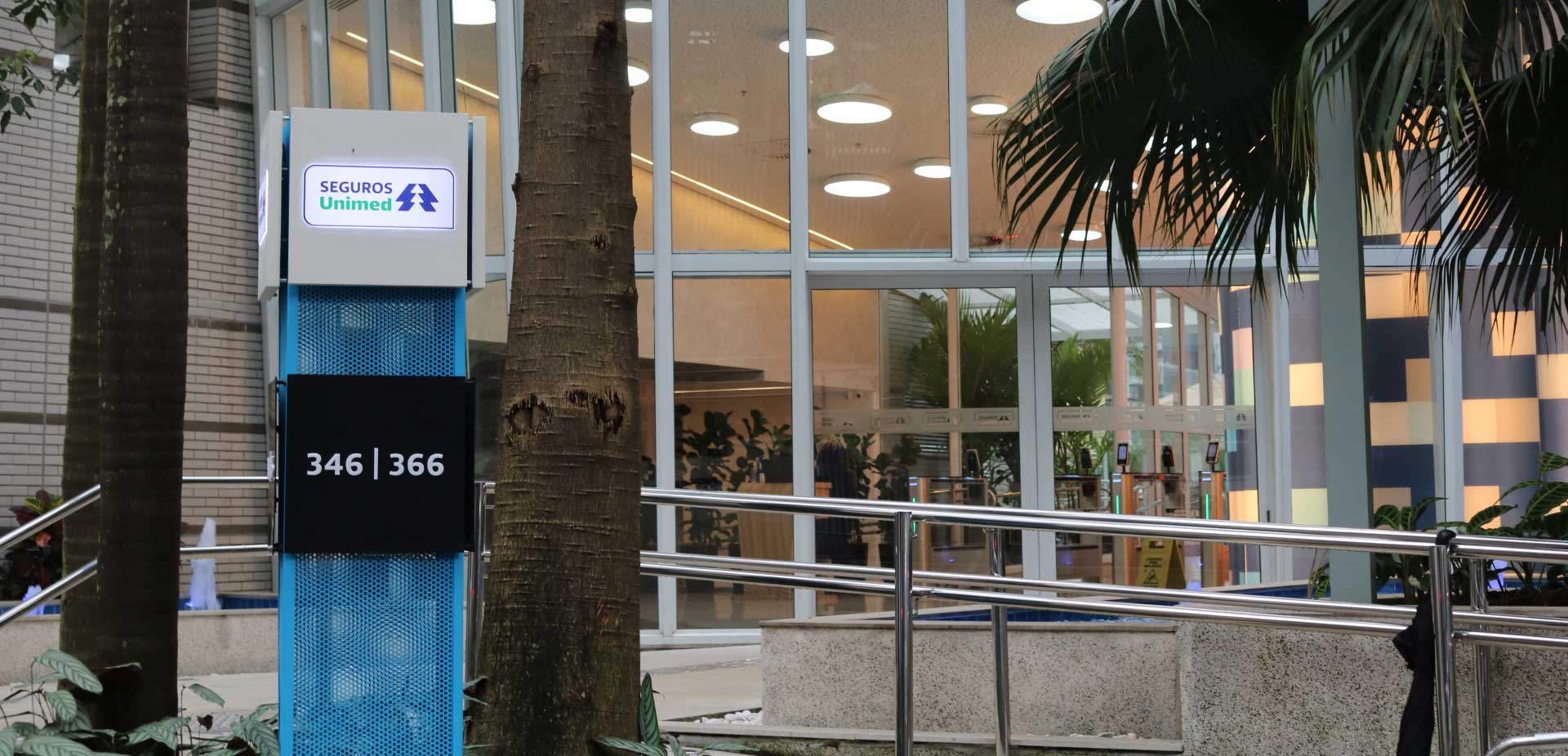Entrance to a modern office building with glass doors, a blue sign displaying Seguros Unimed and address numbers 346/366, surrounded by palm trees and greenery.
