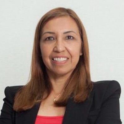 A woman with straight, shoulder-length brown hair smiles at the camera. She is wearing a black blazer over a red top and is posed against a plain, light-colored background.