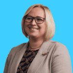 A smiling woman with blonde hair and glasses wears a light-colored blazer over a patterned top, standing against a bright blue background.