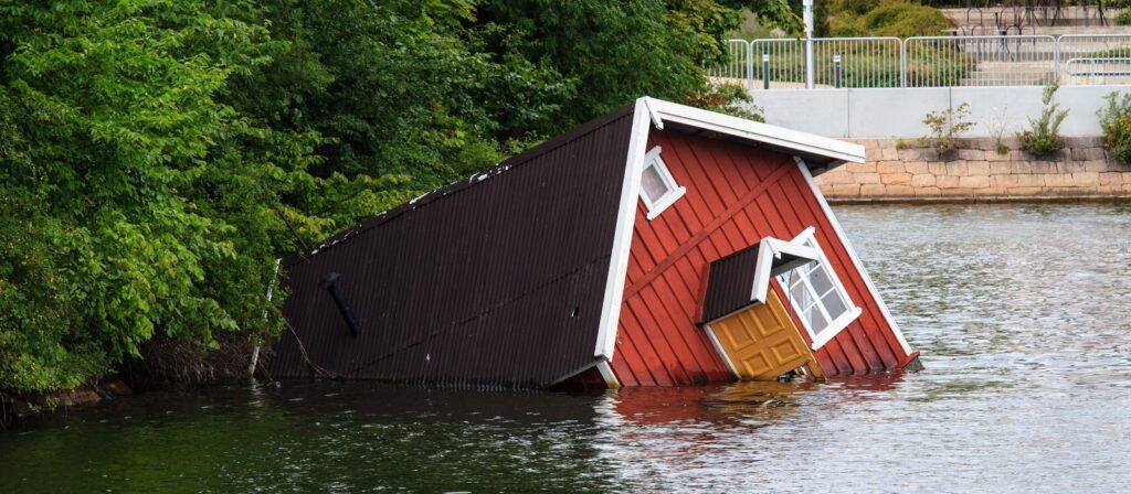 A red house with white trim is partially submerged and tilted in water near a shore, surrounded by green trees. The door and one window are visible above the waterline.