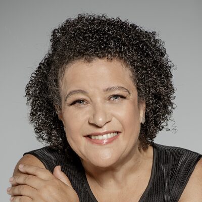 A smiling woman with short, curly dark hair and light skin is wearing a sleeveless black top. She is facing the camera and resting her arm on a surface, with a neutral gray background.