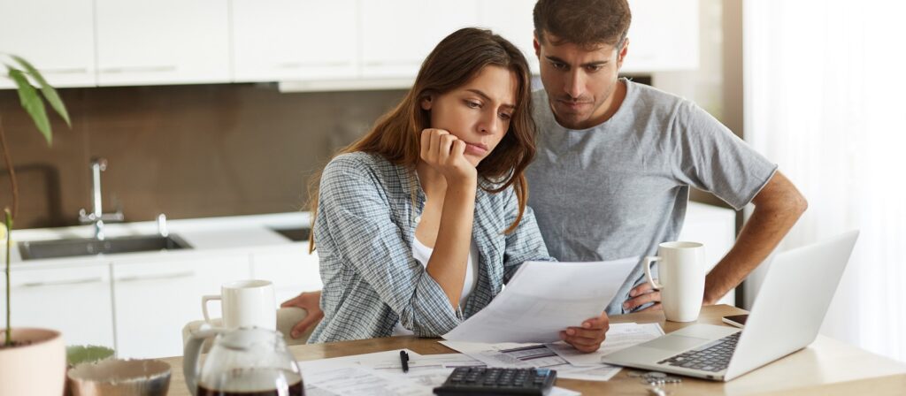 A woman and man sit at a kitchen table, looking concerned as they review documents together. Papers, a calculator, coffee, and a laptop are spread out on the table, suggesting they are managing finances.