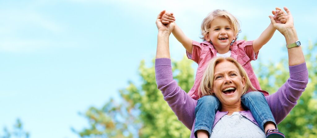 A smiling older woman carries a happy young child on her shoulders outdoors, both raising their hands joyfully against a background of green trees and a blue sky.