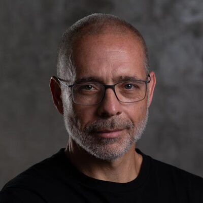 A middle-aged man with a shaved head, gray beard, and glasses wears a black shirt and looks at the camera against a dark, plain background.