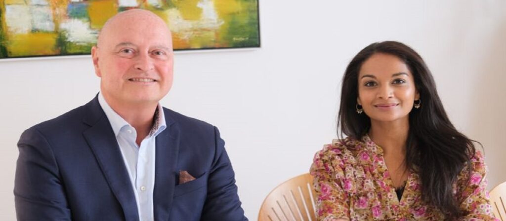 A man in a blue suit and a woman in a floral blouse sit next to each other, smiling at the camera. An abstract painting hangs on the white wall behind them.