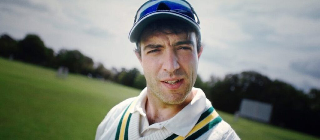 A man wearing a cricket uniform, cap, and sunglasses on his head stands on a grassy field, looking slightly to the side. Trees and a scoreboard are visible in the blurred background under a cloudy sky.