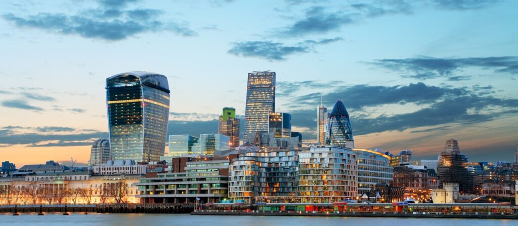 Modern London city skyline at dusk, featuring iconic skyscrapers such as the Walkie-Talkie, the Gherkin, and other buildings, with the River Thames in the foreground and a partly cloudy sky above.