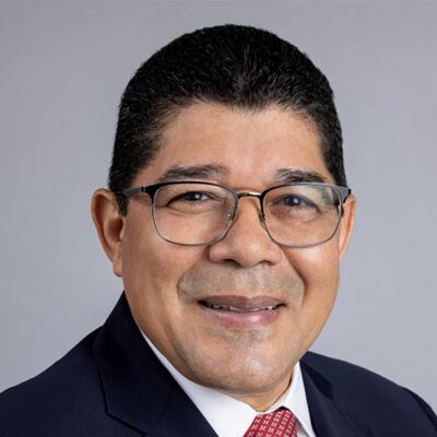 A man with short dark hair, glasses, and a dark suit with a red patterned tie, smiling at the camera against a plain light gray background.