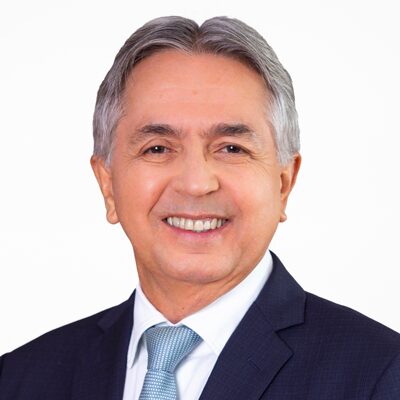 A smiling older man with gray hair, wearing a dark suit, white shirt, and light blue tie, poses in front of a plain white background.