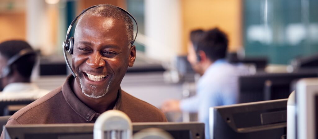 A smiling man wearing a headset sits at a desk in a busy office, working at a computer. Other people with headsets are visible in the background, also working at their desks.