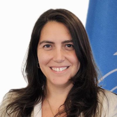 Smiling woman with long dark hair, wearing a light-colored blazer, stands in front of a blue flag with a white emblem on it, against a white background.