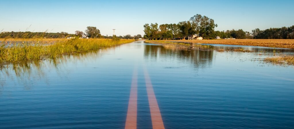 A rural road is flooded, with water covering the pavement and only the faint double yellow center lines visible. Fields and trees border the road under a clear blue sky.