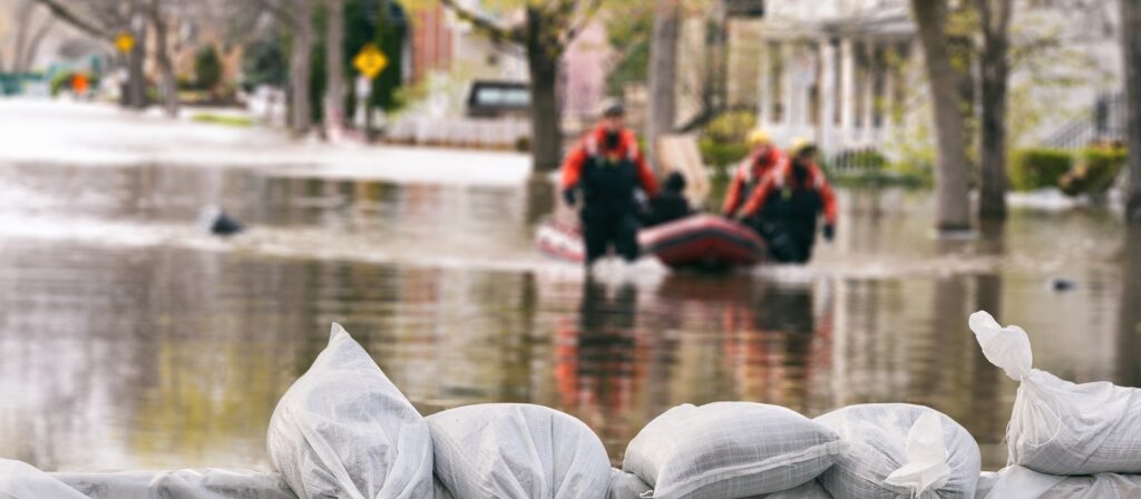Sandbags in the foreground block rising floodwaters, while emergency responders in red suits navigate a rescue boat through a flooded residential street in the blurred background.