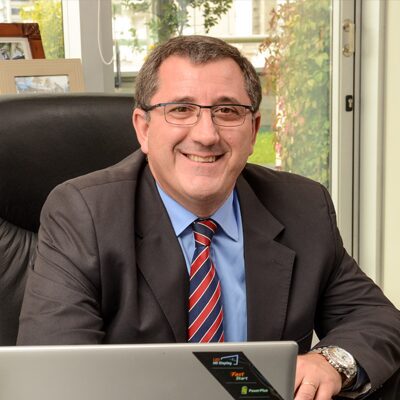 A man wearing glasses, a dark suit, and a striped tie smiles while seated at a desk with a laptop in front of him. There is a window and some greenery in the background.