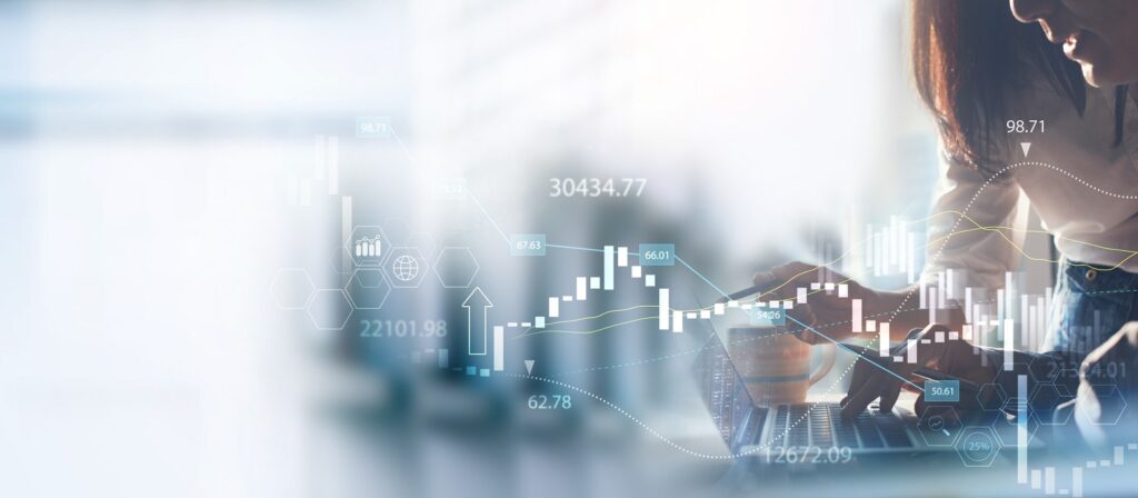 A person works on a laptop with a tablet, overlaid by digital financial graphs, charts, and numerical data, symbolizing analysis, trading, or financial technology in a modern business setting.