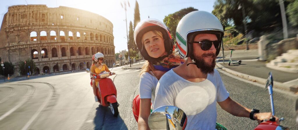 A couple rides a scooter in front of the Colosseum in Rome, Italy, with another person following on a red scooter. The sun is bright and the street is mostly empty.