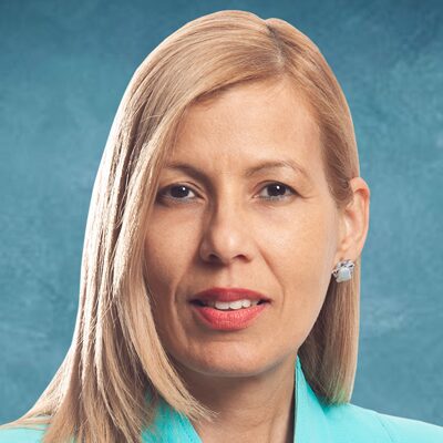 A woman with straight, blonde hair and light skin, wearing a turquoise blazer and silver earrings, poses in front of a blue textured background. She is looking directly at the camera with a neutral expression.