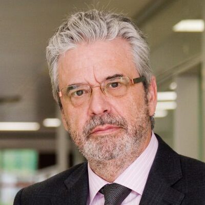 A middle-aged man with gray hair, glasses, and a beard, wearing a suit and tie, stands indoors in a well-lit office setting, looking directly at the camera with a serious expression.