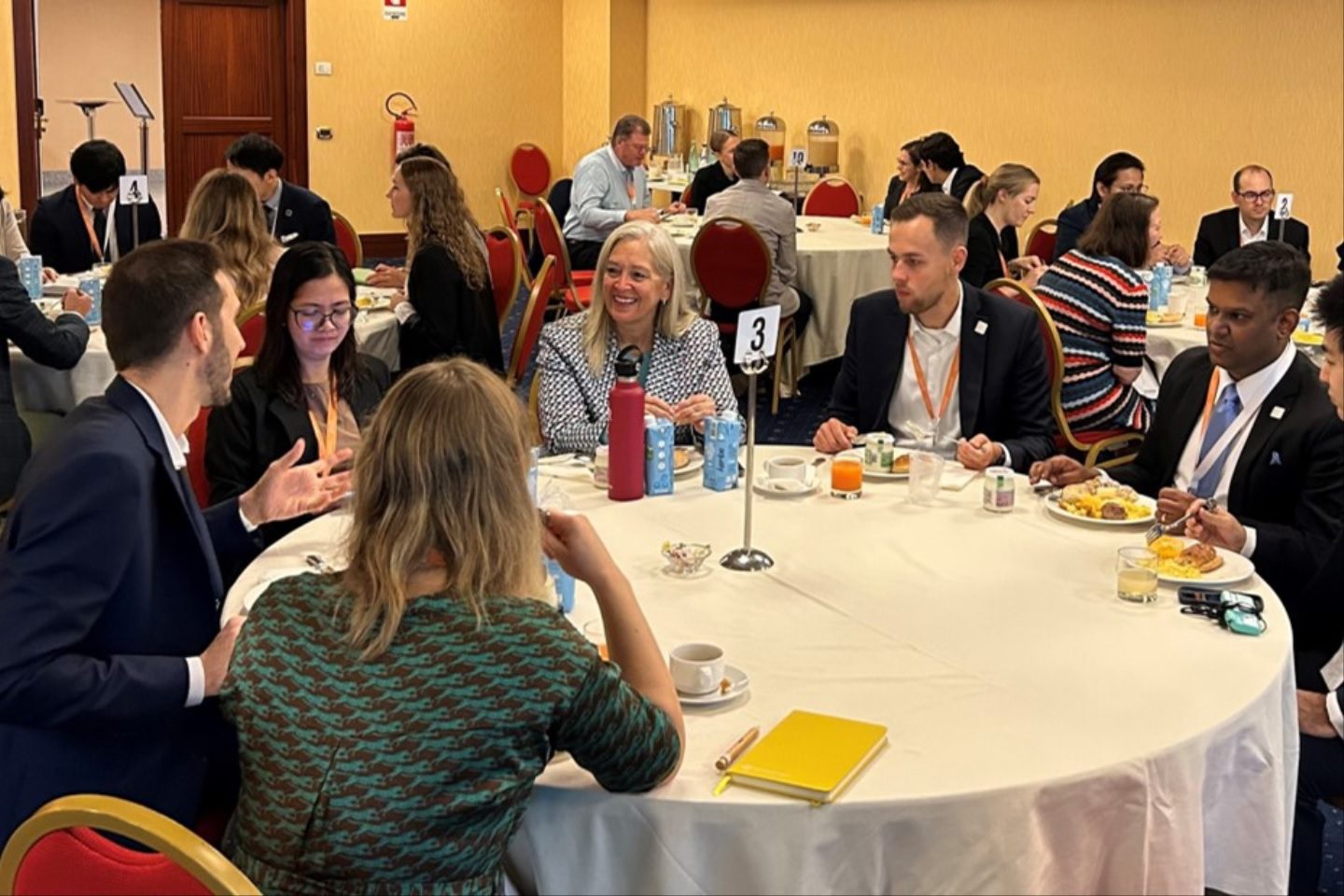 A group of people in business attire sit around a round table at a conference or meeting, engaged in discussion. Notebooks, drinks, and plates of breakfast food are on the table. More people sit at other tables in the background.