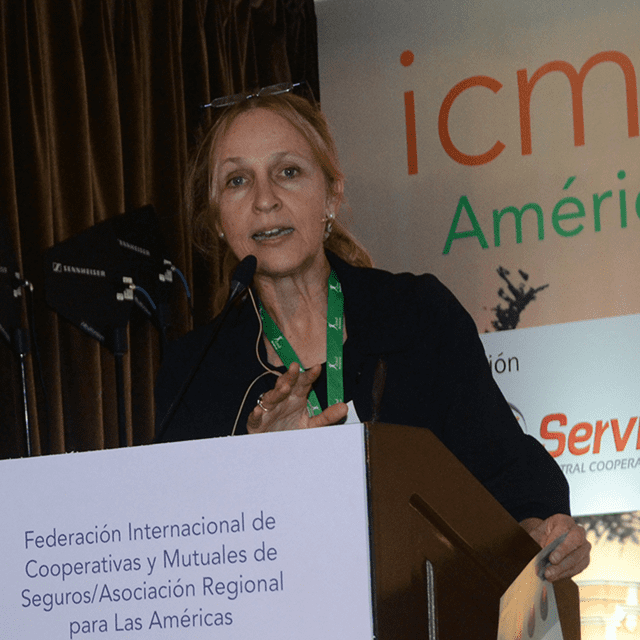 A woman speaks at a podium with a microphone, wearing a green lanyard. Behind her is a banner with Spanish text and the acronym “icm”. The podium displays information about a regional insurance cooperatives association.