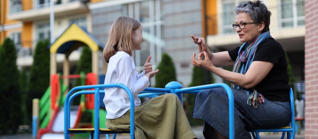 A young girl and an older woman sit on playground equipment outdoors, smiling and communicating using sign language. A colorful play structure is visible in the background.
