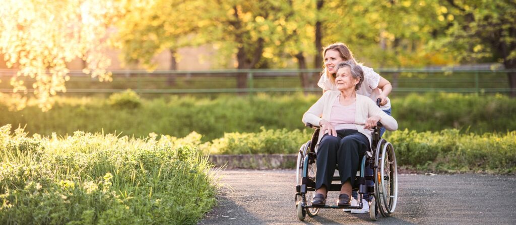 A younger woman pushes an older woman in a wheelchair along a sunny, green park path. Both are smiling, surrounded by lush trees and grass.