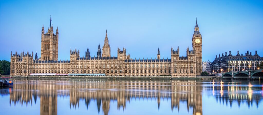 The Houses of Parliament and Big Ben in London are reflected in the calm River Thames at dusk, with a clear blue sky overhead.