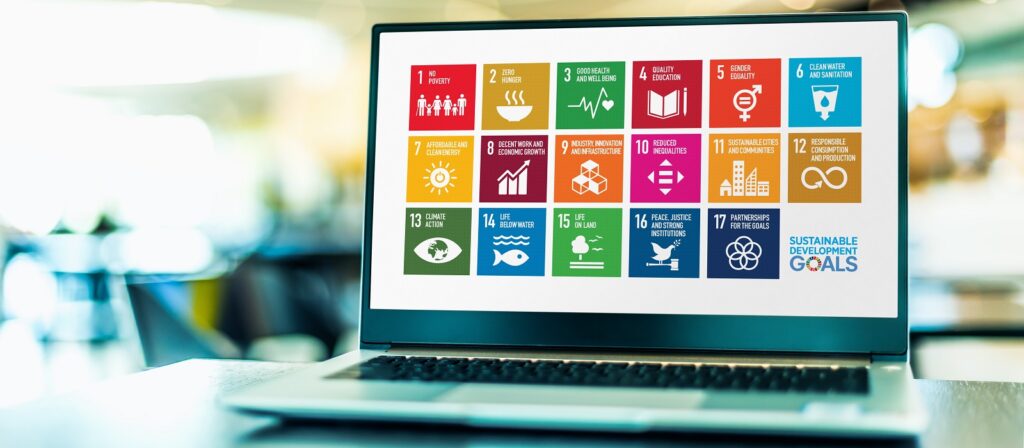 A laptop displays colorful icons and text representing the 17 United Nations Sustainable Development Goals on its screen, set against a blurred office background.