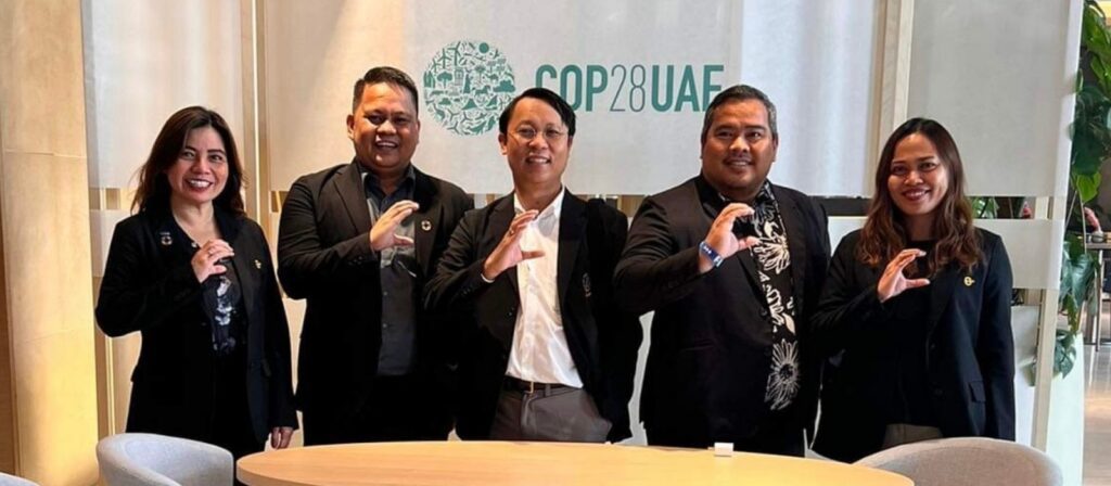 Five people stand in front of a COP28 UAE sign, smiling and making a C shape with their hands. They are dressed in black suits and standing behind a round table in a well-lit room.