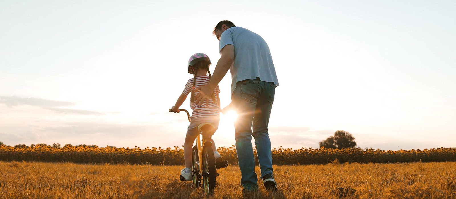 Caring father helps little daughter to ride bicycle in evening meadow. Father teaches little girl to ride bicycle in summer park. Father and little daughter train bicycle riding on rural weekend
