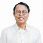 A man with short black hair and glasses is smiling while wearing a traditional off-white Barong Tagalog shirt against a plain white background.