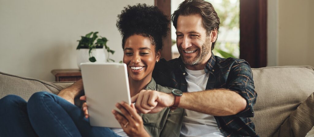 A smiling couple sits on a couch, looking at a tablet together. The woman holds the tablet while the man points at the screen. There’s a plant in the background and they appear relaxed and happy.