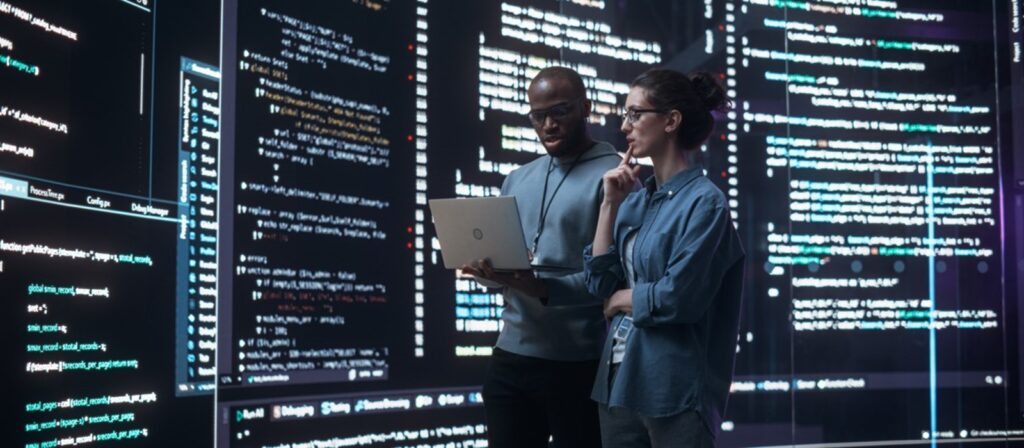Two people stand in front of large screens displaying lines of colorful computer code. One holds a laptop while both analyze the code, suggesting a collaborative programming or cybersecurity environment.