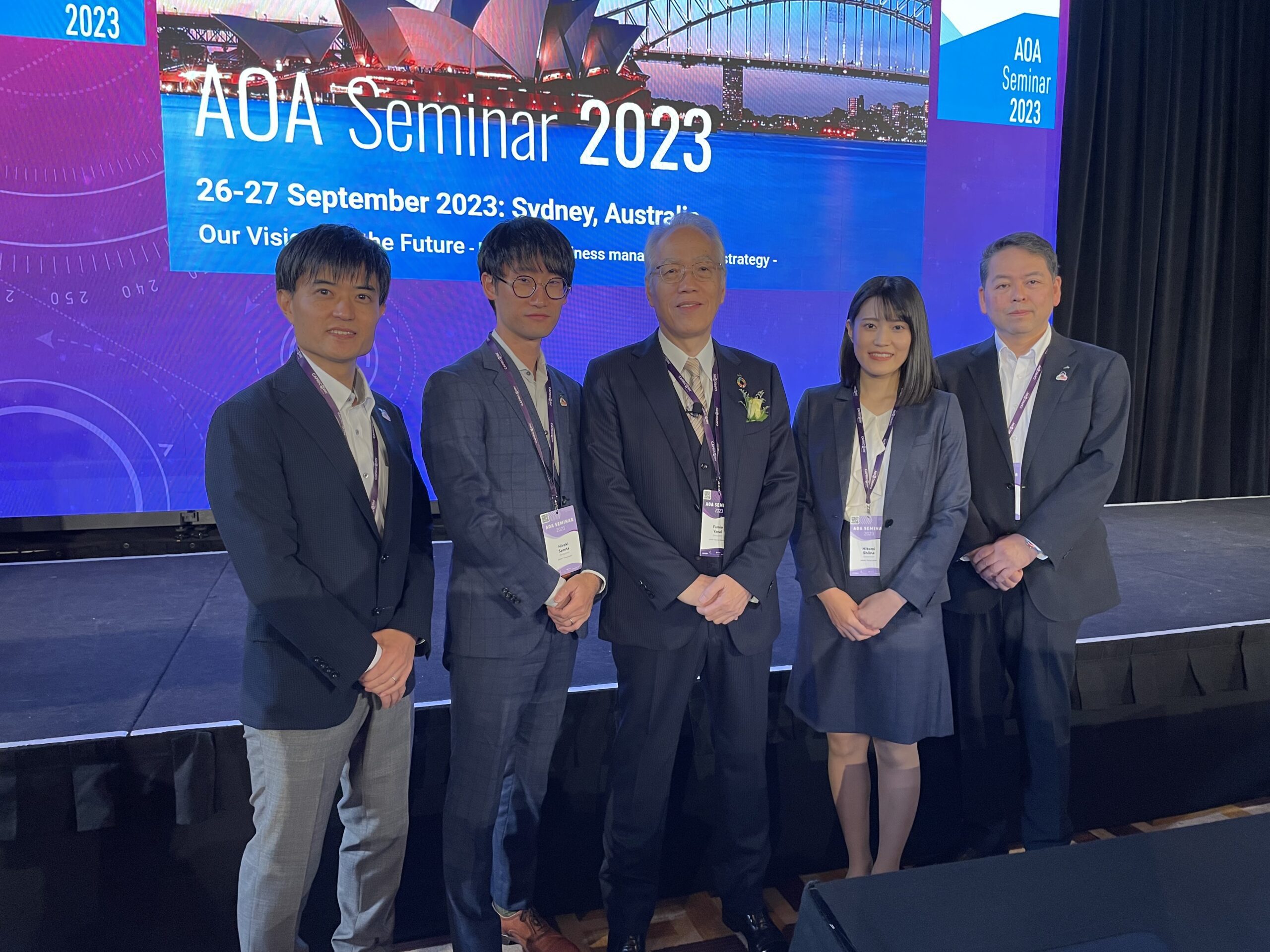Five people in business attire stand together in front of a presentation screen displaying AOA Seminar 2023 with Sydney landmarks pictured, at a professional event in Sydney, Australia.