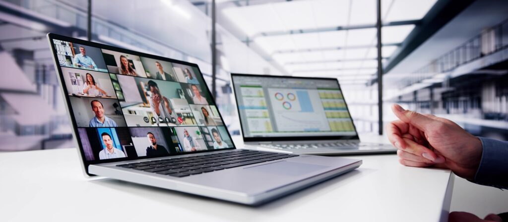 A person sits at a desk with two laptops; one displays a video conference with many participants, while the other shows charts and data, set in a bright modern office space.