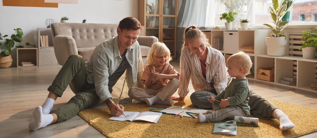 A family of four, two adults and two young children, sit on the floor of a cozy living room, drawing and coloring together. Art supplies are scattered on a yellow rug, and sunlight brightens the space filled with plants.