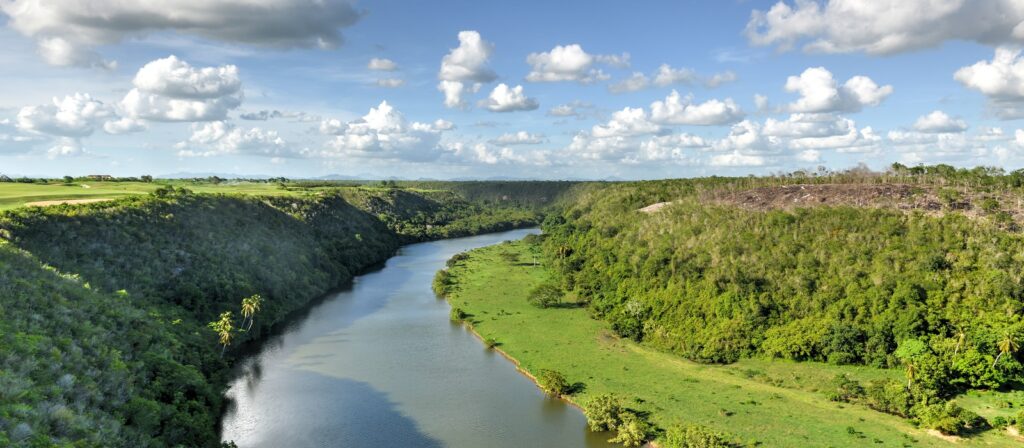 A wide river winds between green, tree-covered hills under a bright blue sky with scattered clouds. The landscape is lush and peaceful, with grassy banks and dense vegetation on both sides.