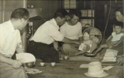 A black-and-white photo showing a group of adults and a child sitting on the floor around a low table indoors, appearing to share a meal or tea. One man serves food while others watch. A hat lies in the foreground.