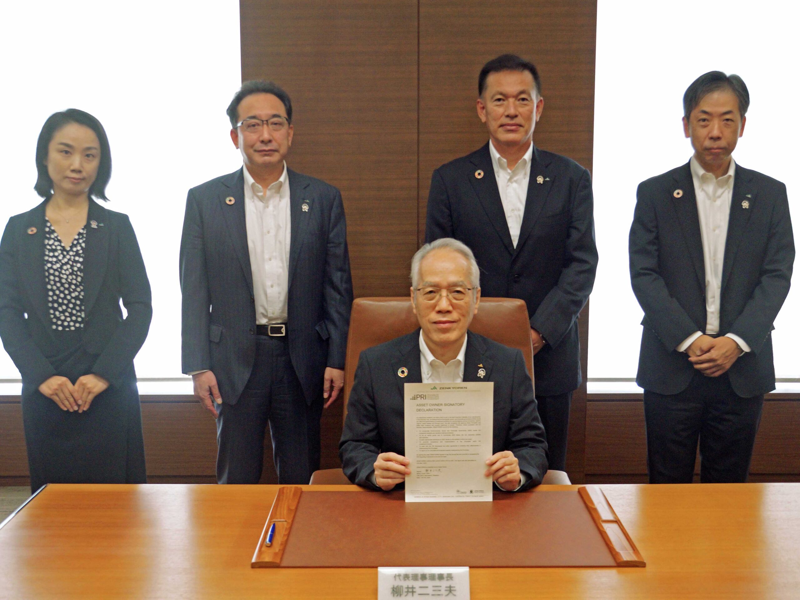 Five formally dressed people pose indoors; four stand behind a seated man holding a document at a wooden desk. The group is in an office setting with large windows in the background.