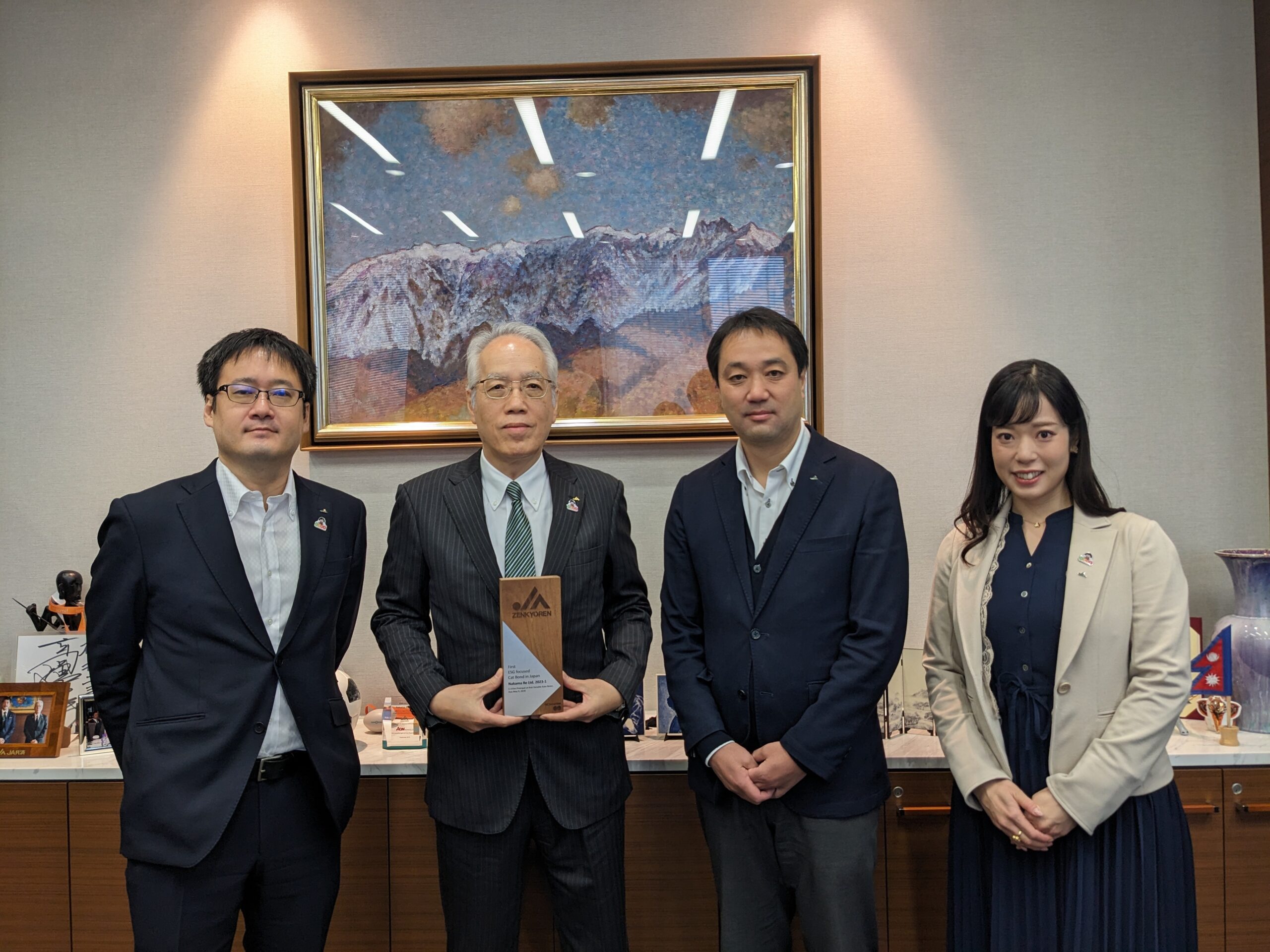 Four people in business attire stand indoors, with the man second from the left holding a plaque. Behind them is a large framed picture of mountains and various items displayed on a shelf.