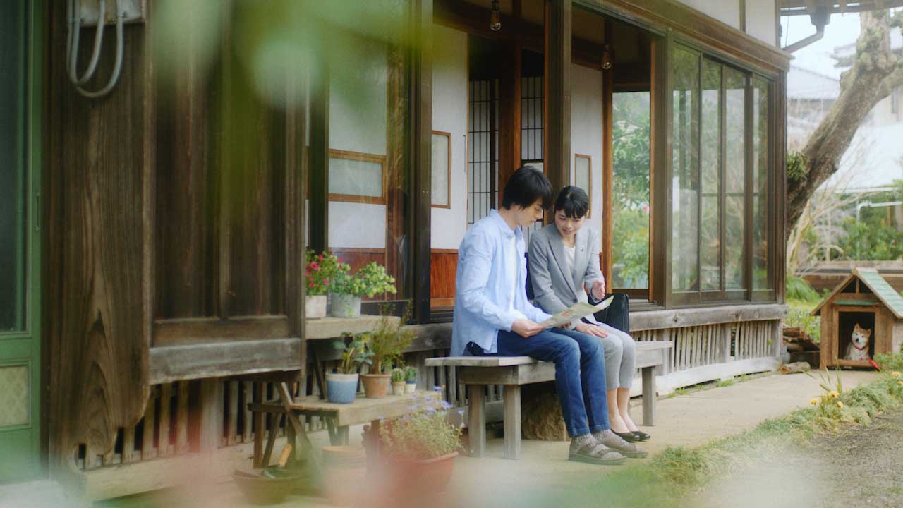 A man and woman sit together on a wooden bench outside a traditional Japanese house, looking at papers. Plants and potted flowers line the porch, and a dog sits in a nearby wooden doghouse.