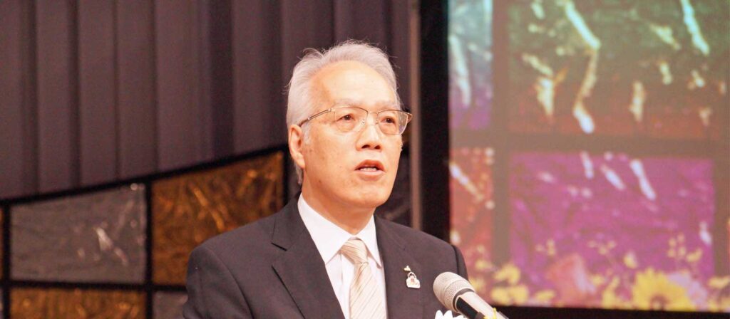 An older man in a suit and tie speaks into a microphone. He has gray hair and wears glasses. Behind him is a colorful, abstract background and dark curtains.