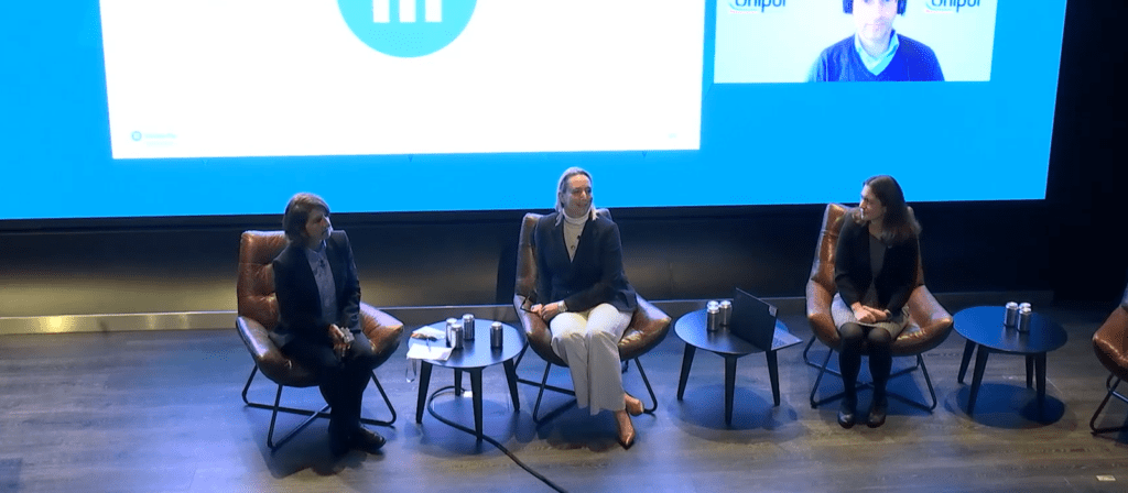 Three people sit on stage in armchairs during a panel discussion. A large screen behind them displays a blue graphic and a video call participant. Small tables with water bottles are positioned beside the chairs.